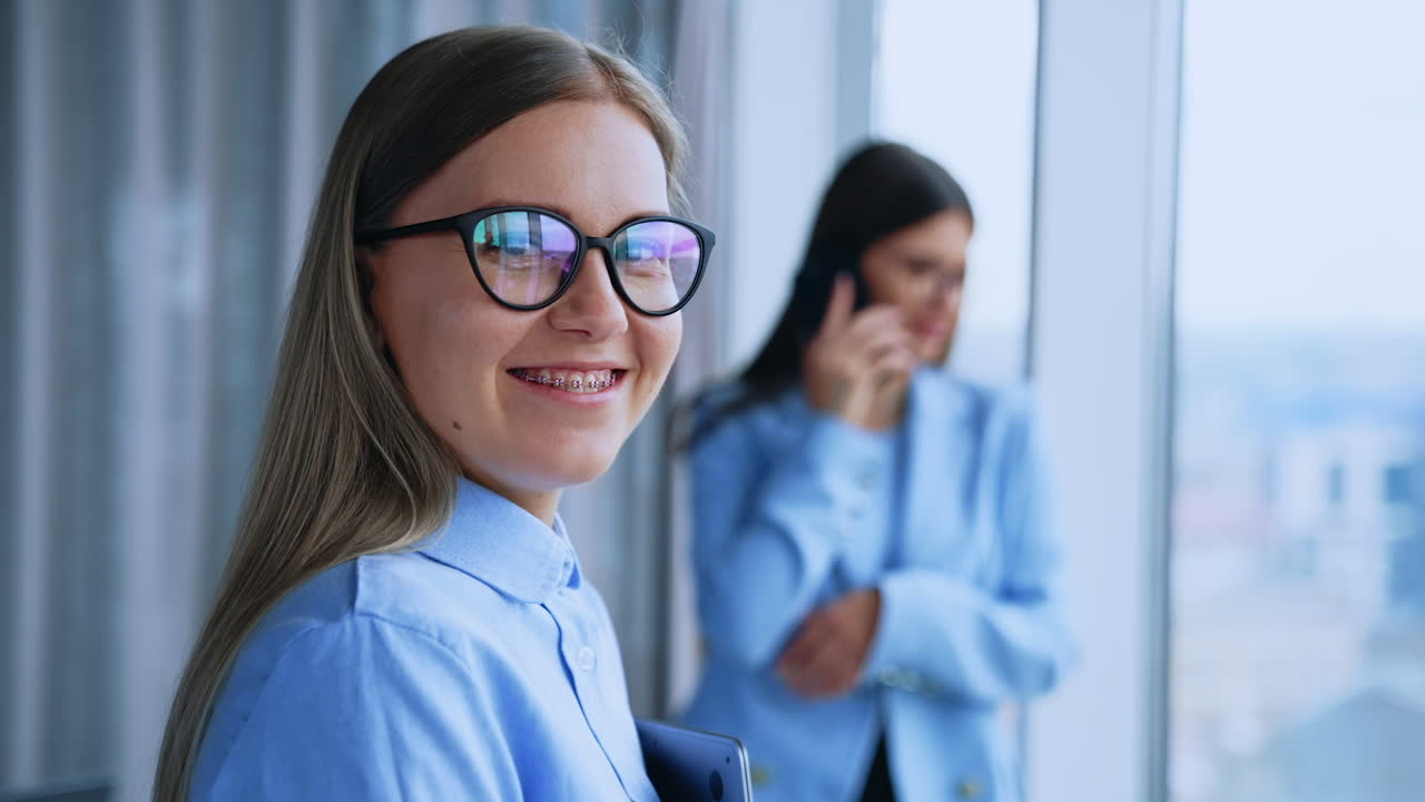 Portrait of a smiling happy young female office employee. Close up. Dark-haired woman speaking on the phone near window at backdrop in blur.