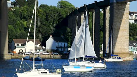 a tall sailboat sails under the tamar and royal albert bridge between devon and cornwall