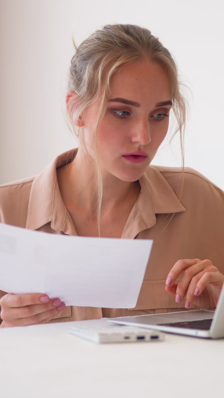Young blonde woman does paper work looking at screen of modern laptop standing on white stylish table in light home office room slow motion