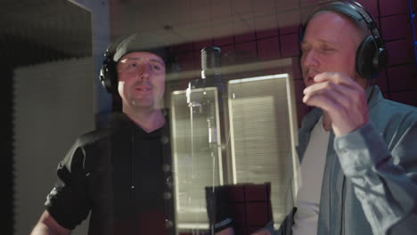 two men in a red soundproof room sing with deep emotion, using expressive hand movements. one wears a black cap and shirt, while the other is dressed in a blue shirt and white inner shirt