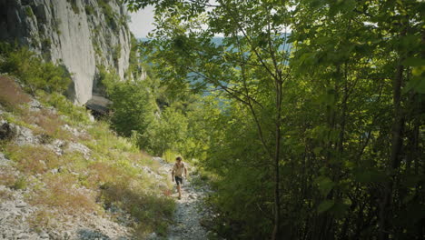 hiker in summer clothes and a backpack walking up a rocky path at the edge of a green forest and a rock clif on the other side