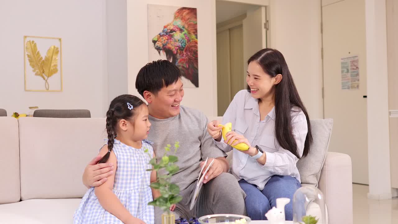 Smiling parents and child enjoy playful banana moment on sofa in bright, cozy living room