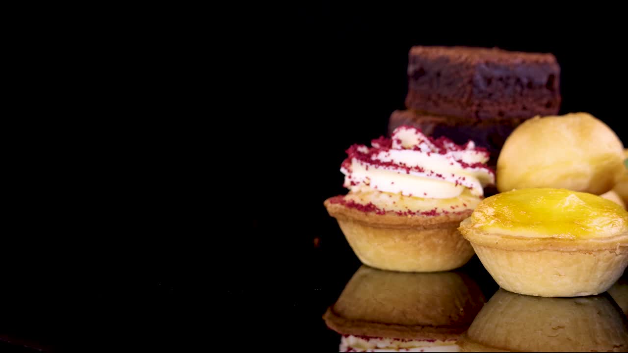 A selection of desserts including brownies, tarts, cream puffs, and strawberries rotates smoothly on a glossy black surface with dramatic studio lighting