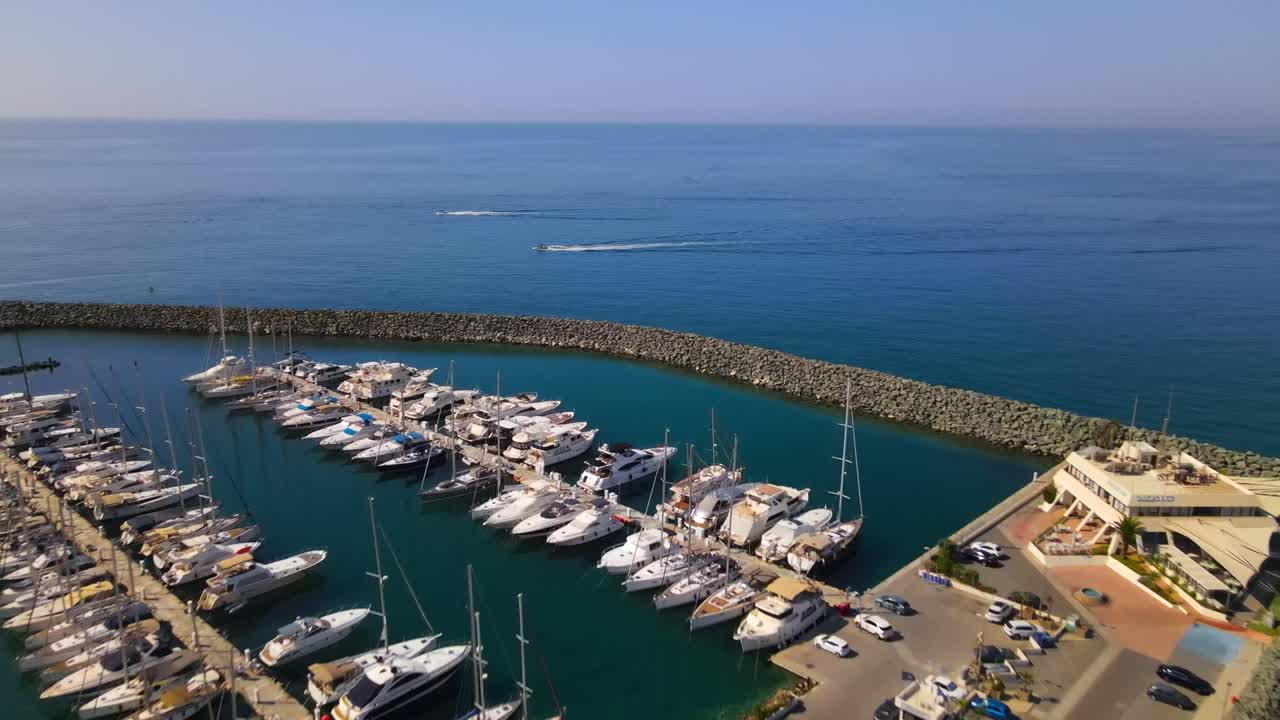 Stunning aerial footage of Sarandë Marina, Albania, showcasing a vibrant coastal scene with yachts, curved breakwater, and serene blue waters under a clear sky, offering tranquility