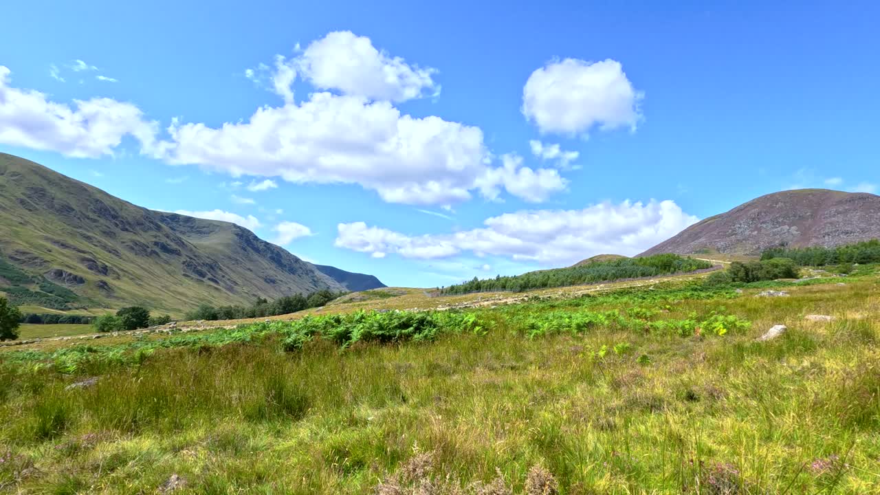 Camera moves steadily along a rugged dirt path through grassy hills under bright daylight, revealing scenic Scottish landscape with blue sky and scattered clouds
