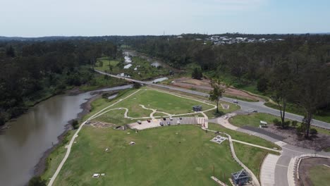 Drone-flying-over-a-recreational-park-towards-a-river-with-bridge-where-cars-are-passing-by