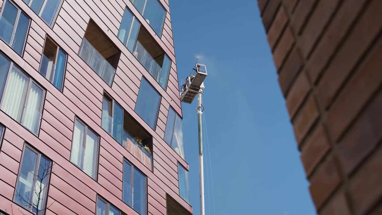 Una piattaforma aerea idraulica solleva gli operai lungo la facciata color rame di un moderno grattacielo sotto un cielo azzurro e limpido, campo lungo