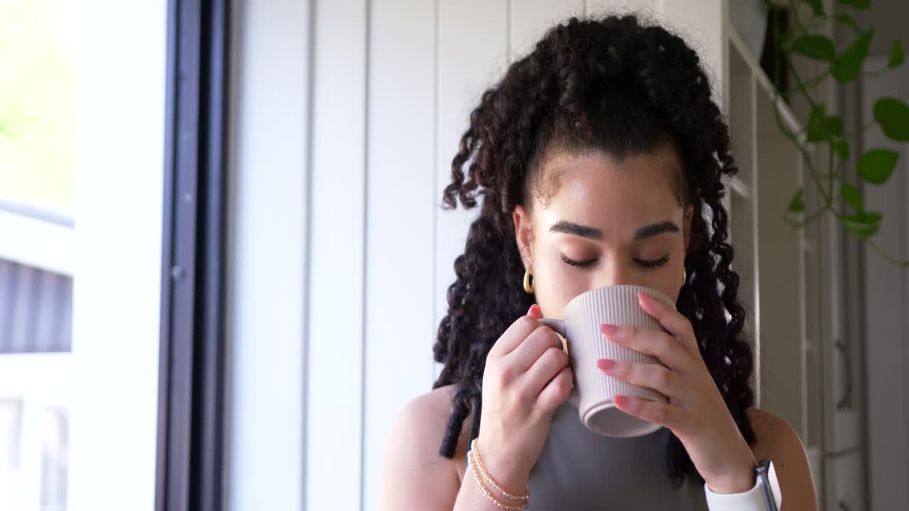Mulher adulta em pé na janela segurando uma caneca canelada, sentindo o aroma e depois sorvendo enquanto observa.