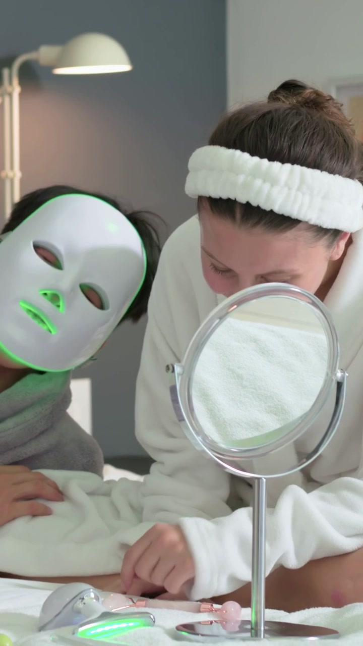 Vertikal video: Kvinde i badekåbe reagerer, mens partner i badekåbe slutter sig til med LED-maske på sengen til hudpleje.