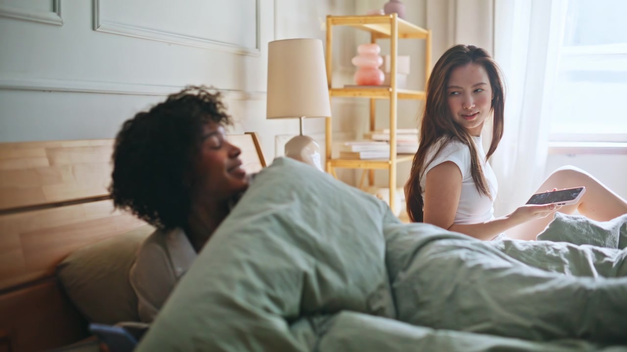 Multiracial girlfriends morning bedroom. Woman using smartphone beside partner.