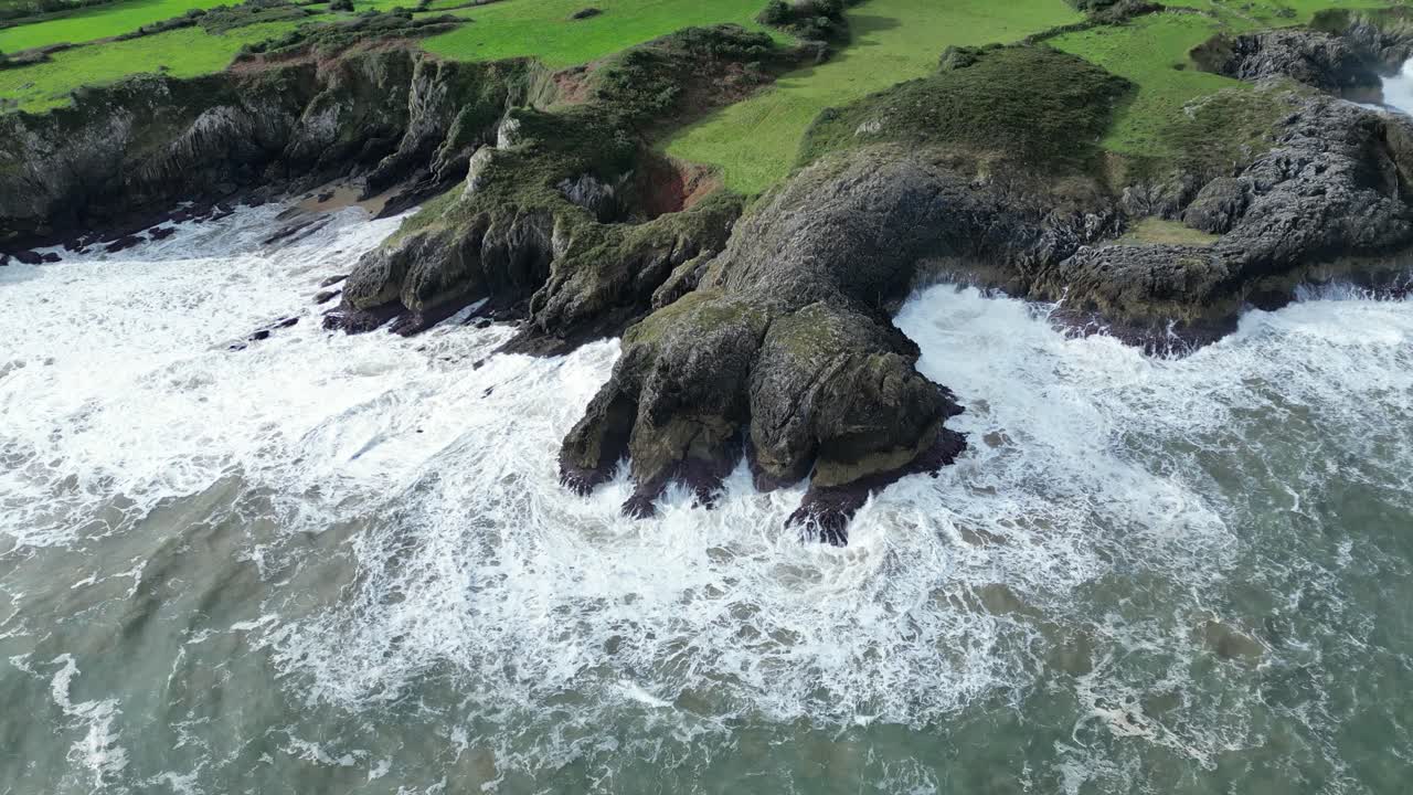 Massive waves crash against a rugged cliffside coastline, flowing through a natural rock arch. In the background, towering mountains create a dramatic scene, showcasing the raw power of the ocean.
