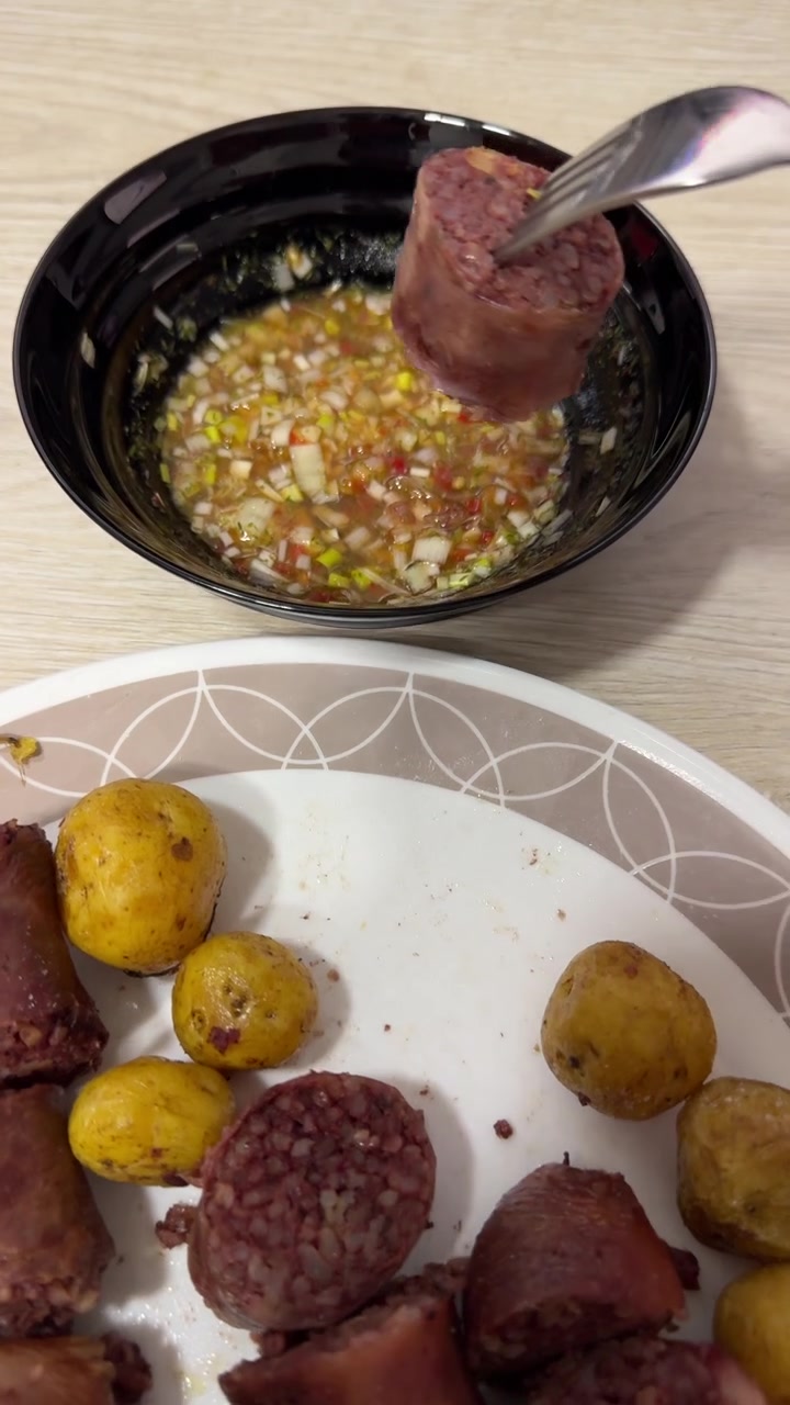 Fatias de morcilla cozida e batatas baby temperadas estão em um prato ao lado de uma tigela de molho aji picado para mergulhar em uma mesa de madeira.
