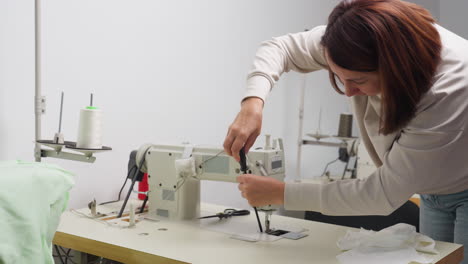 young woman bends over sewing machine using screwdriver to tighten metal part during repair in well-lit studio with white wall background and visible spool of white thread on adjacent stand