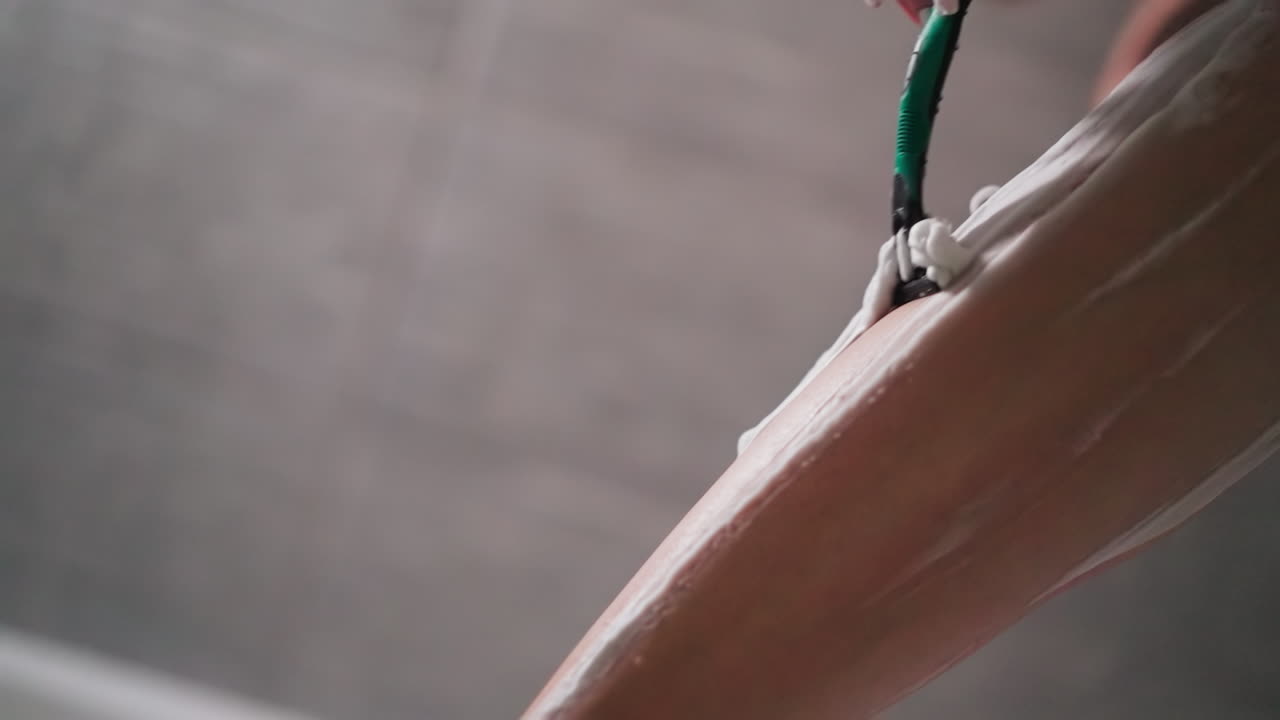 mujer con afeitadora se afeita el pelo de las piernas en la ducha de primer plano. dama elimina el pelo del cuerpo de la piel cubierta con crema en el baño. mujer hace rutina de belleza