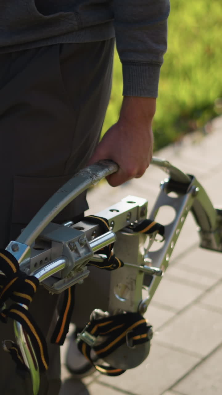 Lower half of person using metal stilts with footrests featuring black straps, wearing dark gray loose-fitting pants and long-sleeved shirt, walking on paved surface with grass background