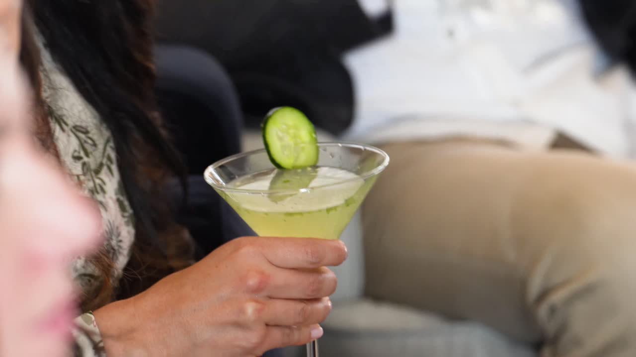 Woman Holds Fancy Cucumber Martini at Cocktail Party with a Group of People seen Speaking in Background and Nodding Foreground. Beverage or Mixed Drink is Green in Color with a Wheel Garnish No Faces
