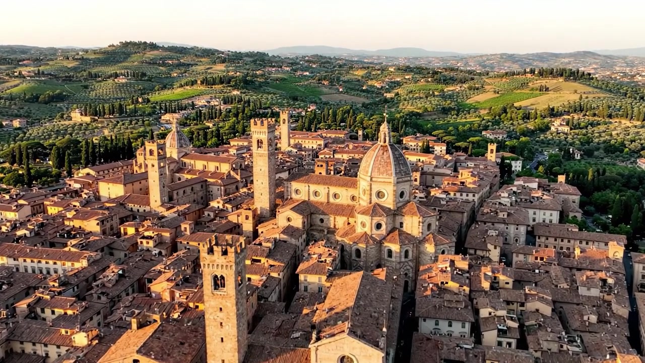 Veduta aerea di una città storica circondata da vivaci colline verdi, che mostra meraviglie architettoniche e paesaggi scenografici baciati dalla calda luce del tramonto.