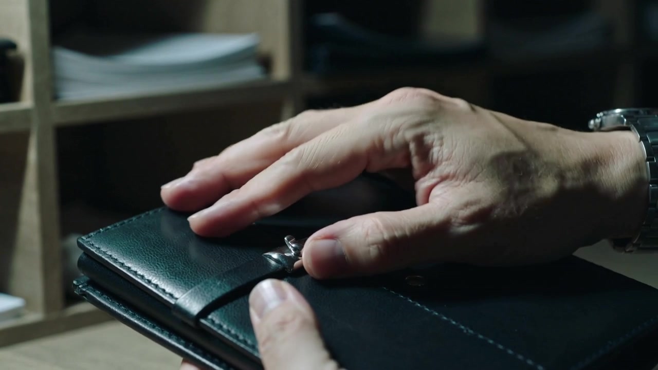 Closing adult hands securing black leather folio on desk, pressing metal clasp with wristwatch