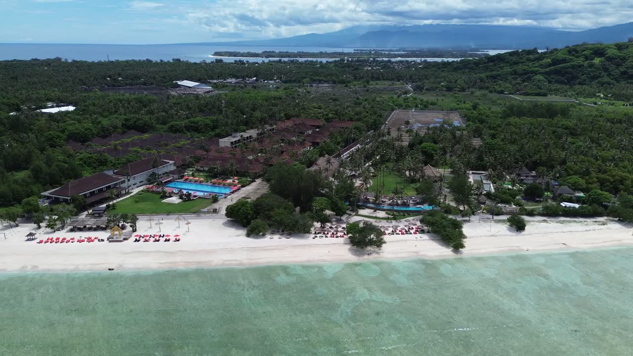 Pan left showing resort buildings, palm trees and sandy beach under a bright sky