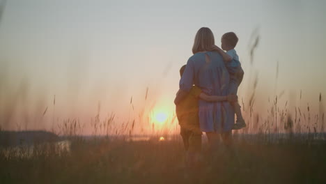 un momento sereno capturando la vista trasera de una madre en un vestido azul sosteniendo a su hijo pequeño mientras el niño mayor está a su lado, todos mirando la puesta de sol sobre un lago tranquilo