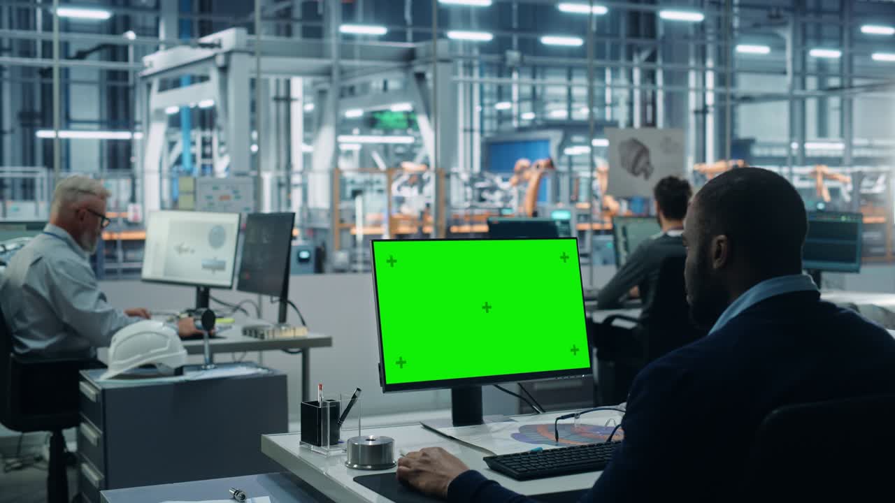 자동차 공장 사무실: 흑인 남성 자동차 수석 엔지니어가 그의 책상에 앉아 녹색 화면 크로마 키 컴퓨터에서 일하고 있습니다. 자동 로 팔 조립 라인 제조. 어깨에서 중간.