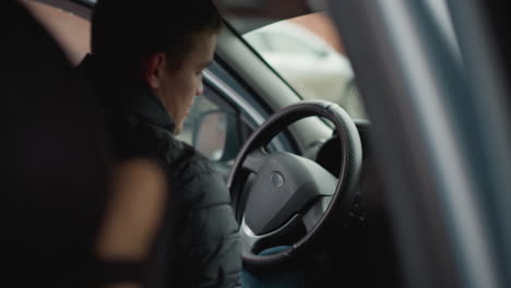 side view of car enthusiast wearing winter jacket entering car sitting on driver seat placing hands on steering wheel preparing for drive in urban parking lot with blurred vehicles in background