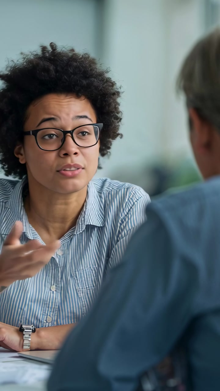 African American woman in striped shirt and coworker gesturing, speaking at desk with docs, glasses