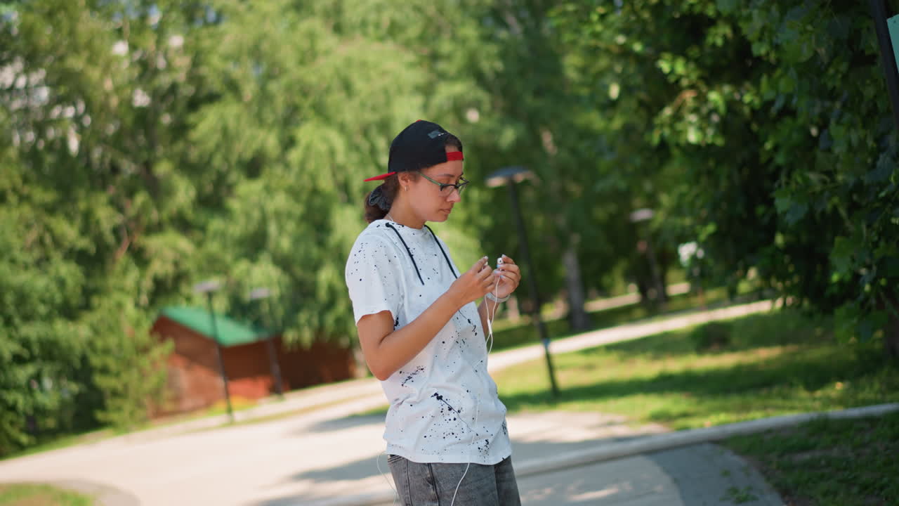 아시아계 십 대 Sunny Park이 휴대폰을 확인한 후 좌절감을 느끼지만 헤드폰을 끼고 미소를 지으며 안도하는 모습, 자연스러운 야외 인물 사진, 늦은 아침 햇살, 푸른 나무, 캐주얼한 모자와 티셔츠