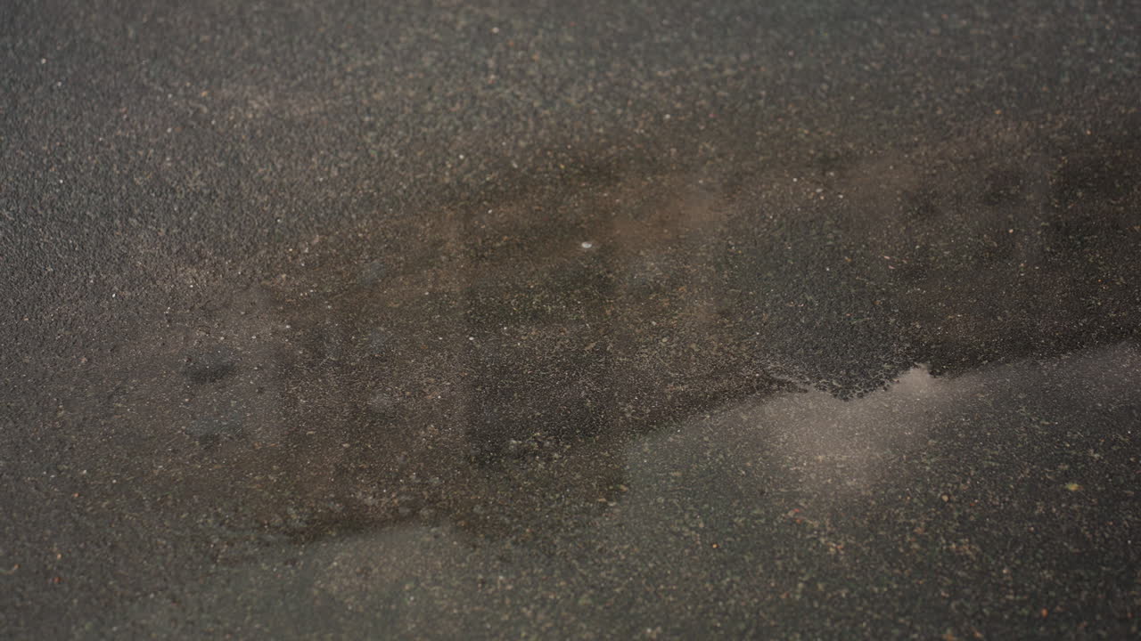 Superfície de asfalto molhado com gotas de chuva, poças brilhantes e pequenos respingos criando uma superfície texturizada, reflexos urbanos suaves e brilho lustroso, atmosfera cinematográfica escura, estudo de movimento em close-up