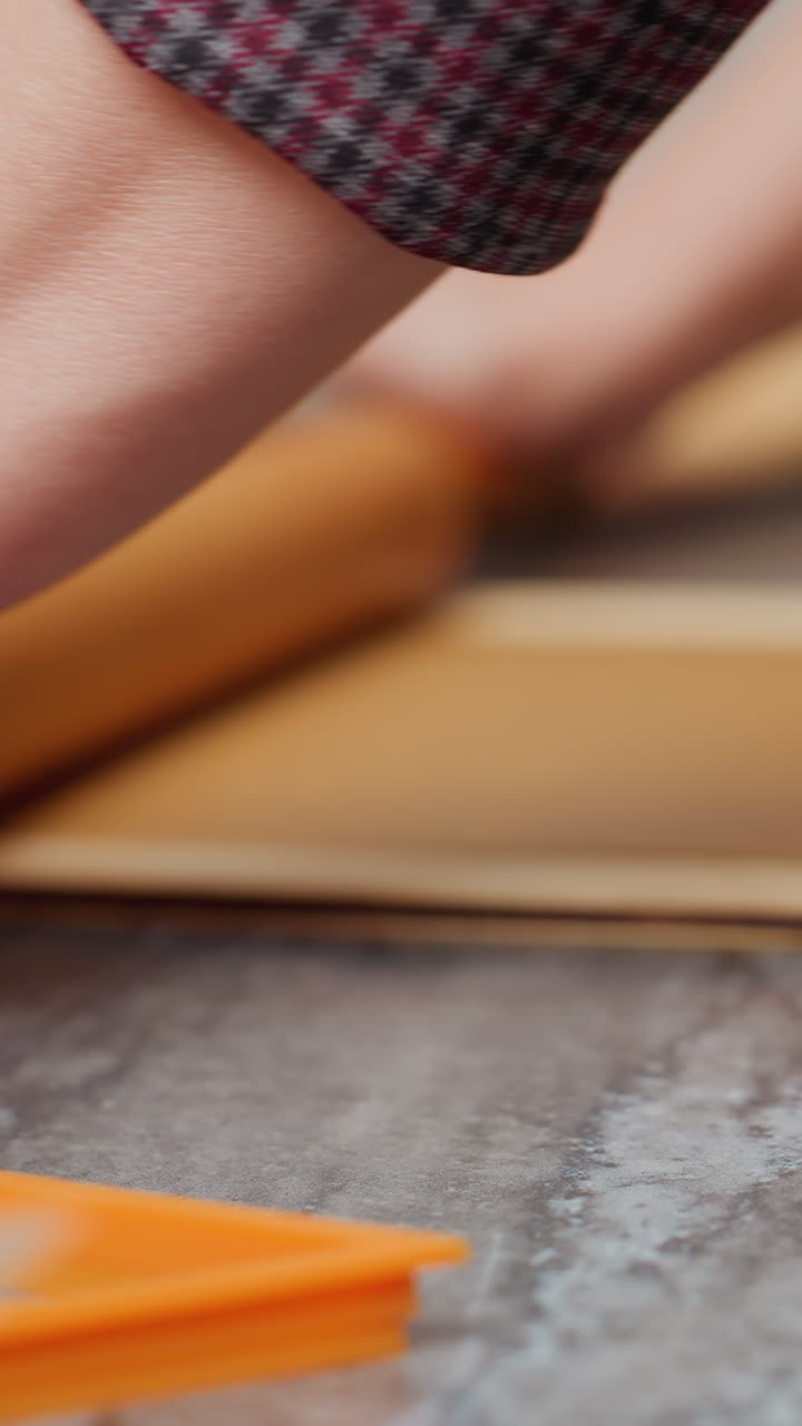 Close-up of woman hands rolling brown dough with a wooden rolling pin, left wrist adorned with a pink beaded bracelet, slight blur in background featuring candle and baking tools