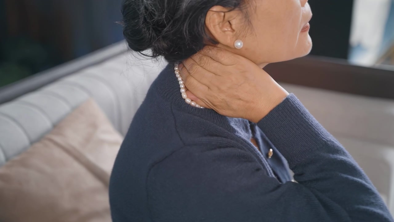 Donna anziana che massaggia le articolazioni del collo con sintomi di dolore dovuti a spondilosi cervicale, osteoartrite, stiramento muscolare del collo, infiammazione, in un contesto di prodotti sanitari, seduta sul divano