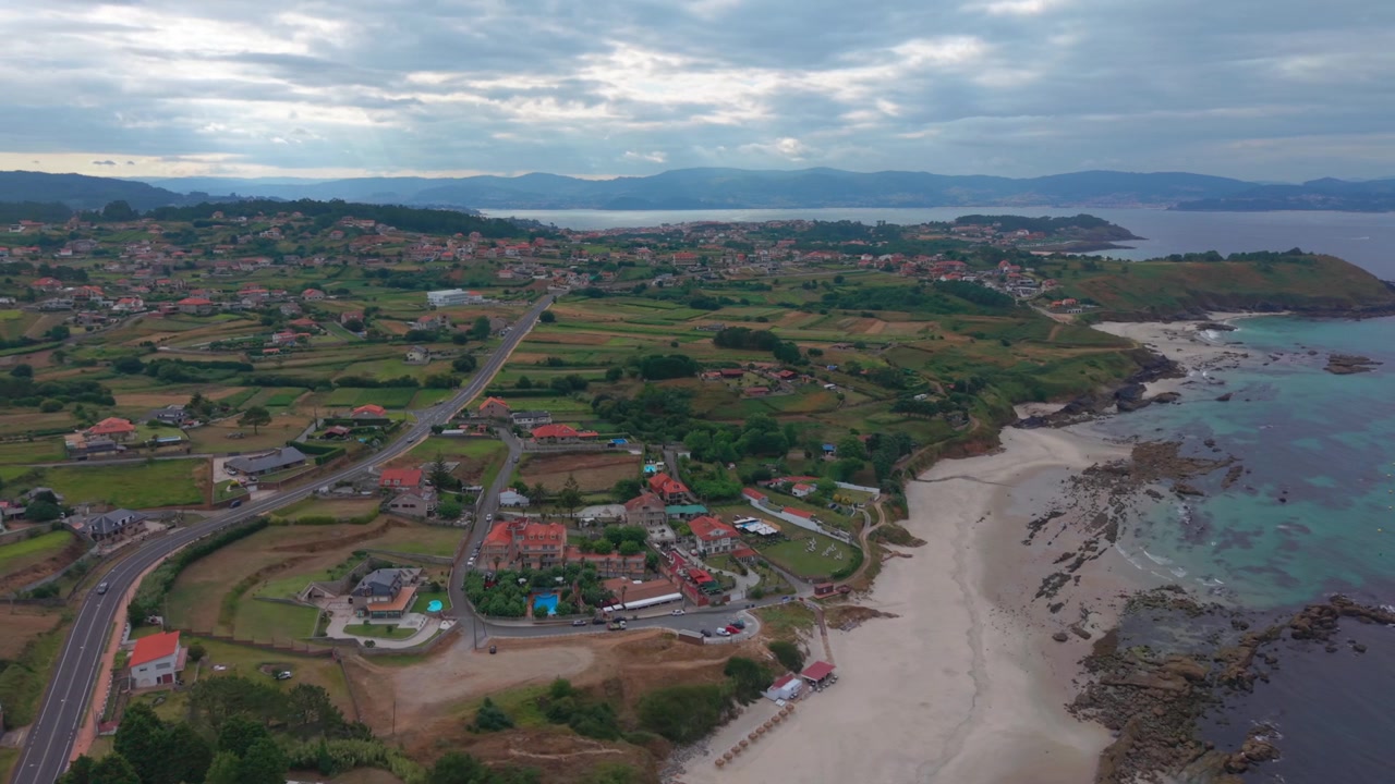 Luftfoto med drone over en kystby og et boligområde ved siden af en sandstrand med klippekyst og turkisblåt vand langs Galiciens kystlinje i Spanien