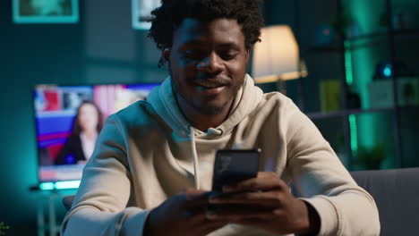 african american man scrolling through social media on smartphone while relaxing on a couch, cozy loft with neon lights. person showcases comfort, online activity and digital engagement. camera a.