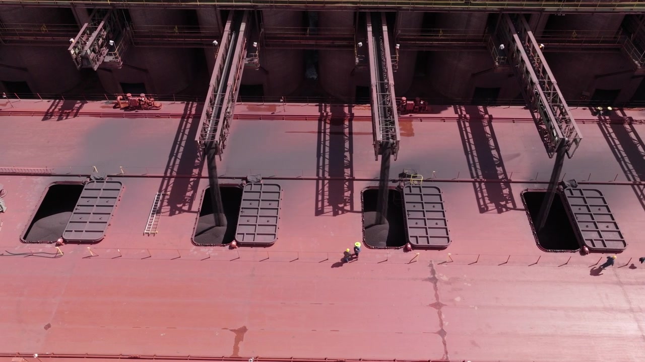 Blue collar workers in PPE supervising natural iron ore resources loading into ship docked in port city