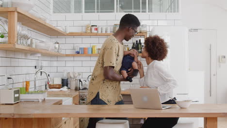 busy family in kitchen at breakfast with mother working on laptop and father caring for baby son