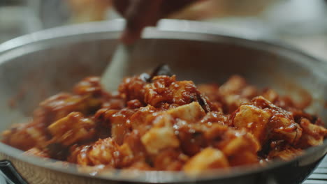 hand of chef stirring spicy tofu dish with spatula in large pan