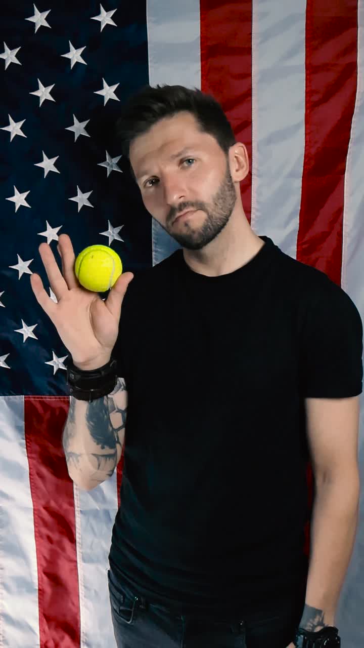 Person with tattoos bouncing a vibrant yellow colored tennis ball in his hand and showing it to the camera in slow motion while standing in front of a vertical hanging American US flag. happy man