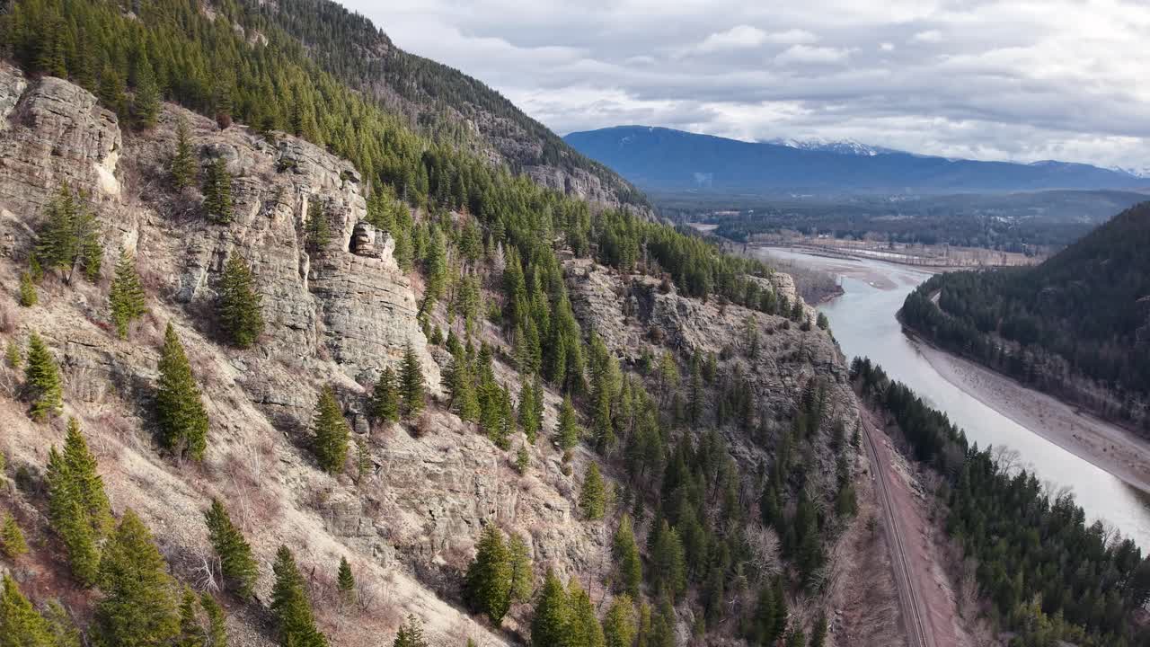 Riprese aeree con drone di una collina rocciosa e boscosa che scende verso un fiume e una strada tortuosi vicino a Whitefish, Montana, con montagne e cielo sullo sfondo