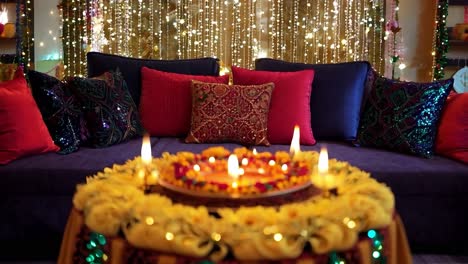 burning candles are standing on a flower arrangement placed on a table in front of a sofa. the room is decorated for diwali festival