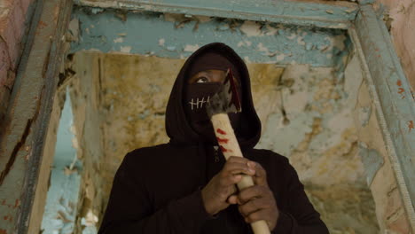 bottom view of man wearing black balaclava and holding an ax in a ruined building 1