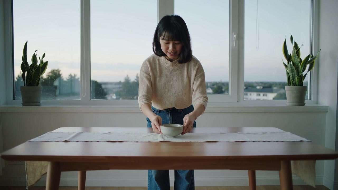 セーターを着た女性が、自宅のダイニングテーブルで不揃いなランナーを整え、陶器のボウルを中央に置いている。