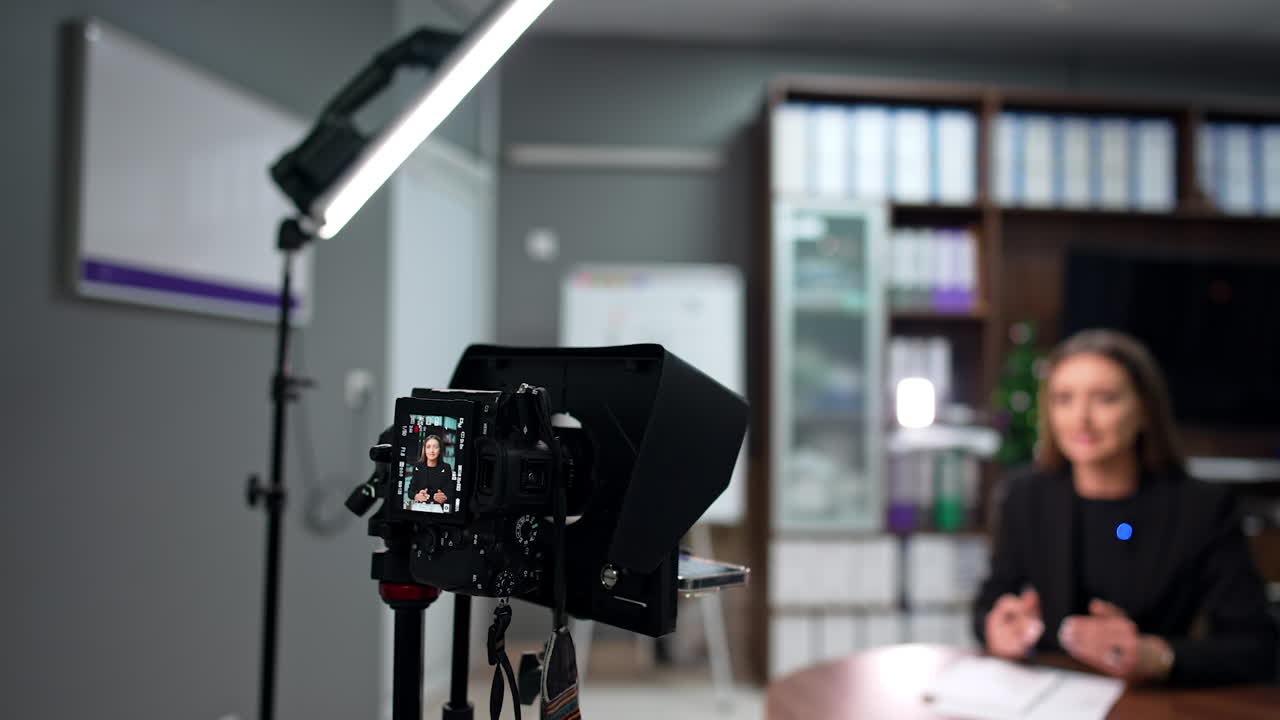 Attractive Caucasian brunette in black jacket records a blog in the office. Camera on tripod display shows the female blogger. Blurred backdrop.