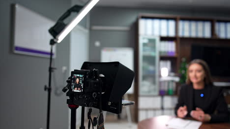 attractive caucasian brunette in black jacket records a blog in the office. camera on tripod display shows the female blogger. blurred backdrop.