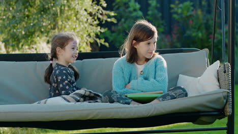 Two-sisters-are-relaxing-on-a-swing-in-the-backyard-of-the-house.-Clear-summer-day