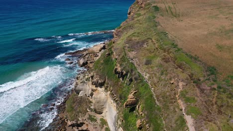 Luftflug-Mit-Blick-über-Eine-Klippe-Im-Meer,-Von-Der-Aus-Man-Eine-Wiese-überblickt-Und-Die-Wellen-Gegen-Den-Felsen-Krachen-Sieht,-Wobei-Verschiedene-Ebenen-Von-Steinen-Und-Verschiedene-Blautöne-Im-Wasser-In-Kantabrien,-Spanien,-Sichtbar-Werden.