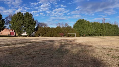 a stationary camera captures an empty sports ground with a soccer goal, dry grass, and scattered trees under bright daylight and partly cloudy skies