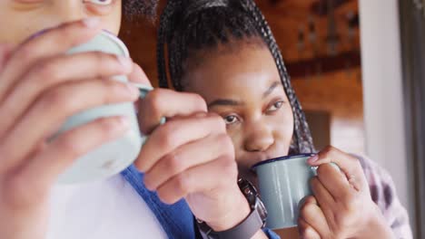 happy african american couple looking through window and drinking coffee in log cabin, slow motion