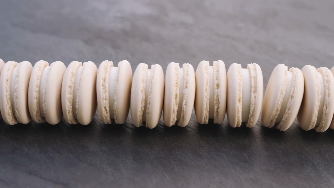 Close-up view of pale cream macarons arranged in continuous horizontal row on matte dark slate, gently shifting with subtle motion, soft light, smooth texture, refined neutral styling