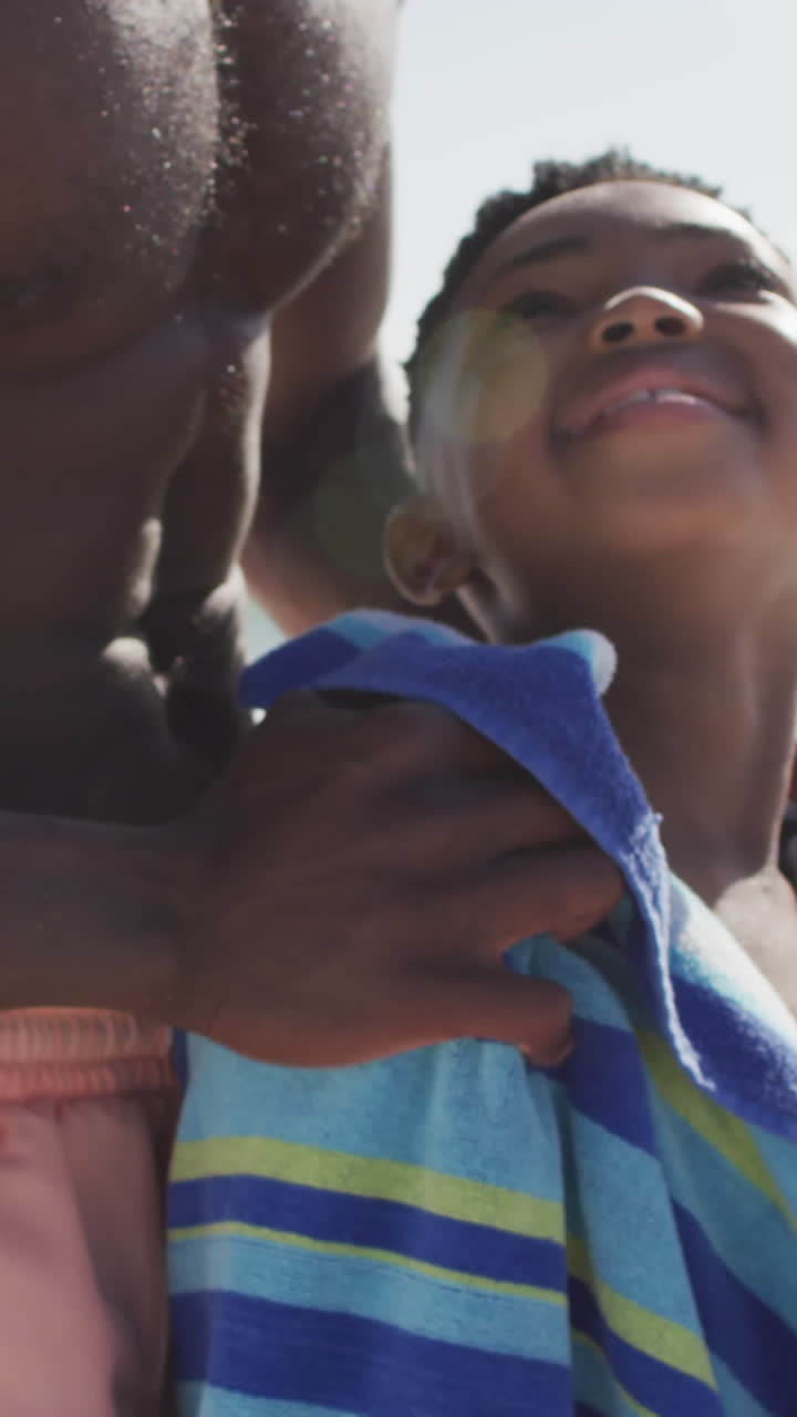 sonriente padre afroamericano toallando a su hijo en una playa soleada