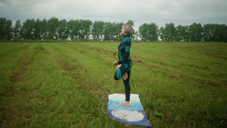 side view of middle-aged woman standing with her right leg on colorful yoga mat in tree pose, practicing yoga in a vast grassy field with trees lining the horizon